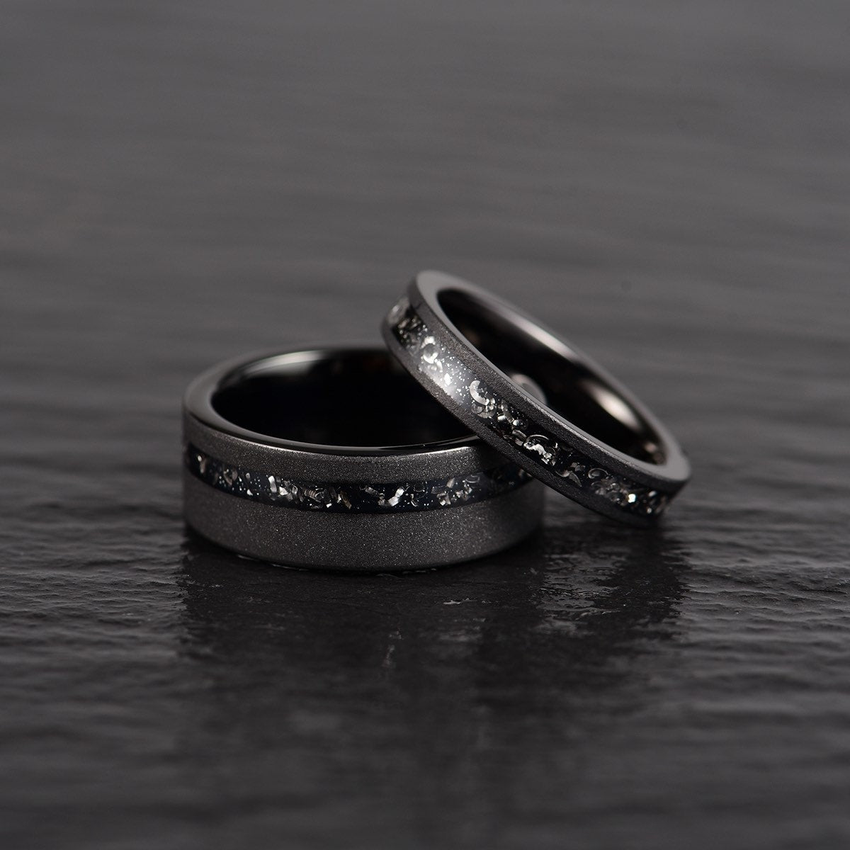 Two crushed meteorite inlay sandblasted black tungsten men's wedding band laid down on top of eachother